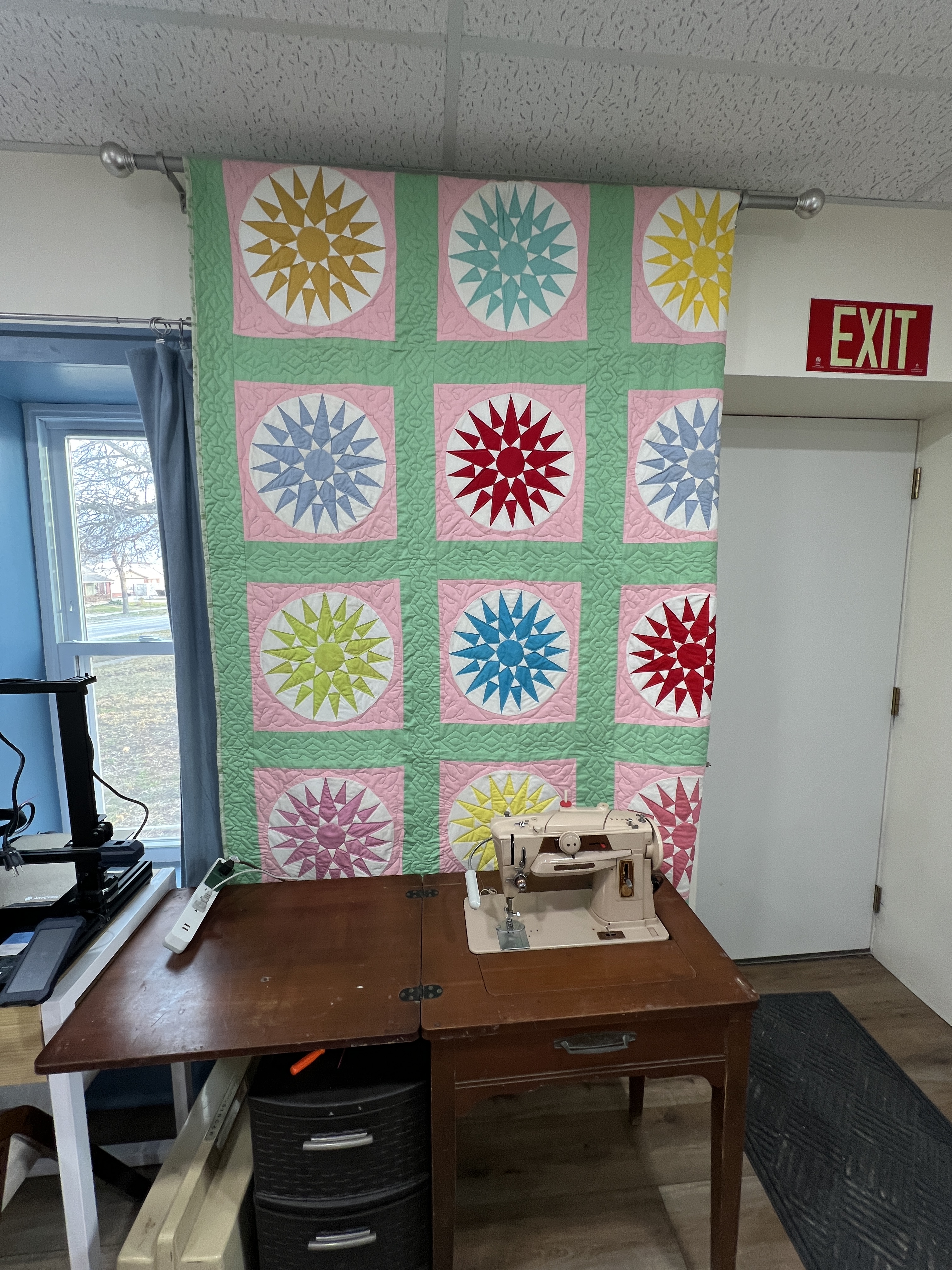 A Singer 401a sewing machine in a brown cabinet sitting in front of a handmade quilt hanging from a rod near the ceiling.  The quilt has many colors but is predominantly pink and green.
